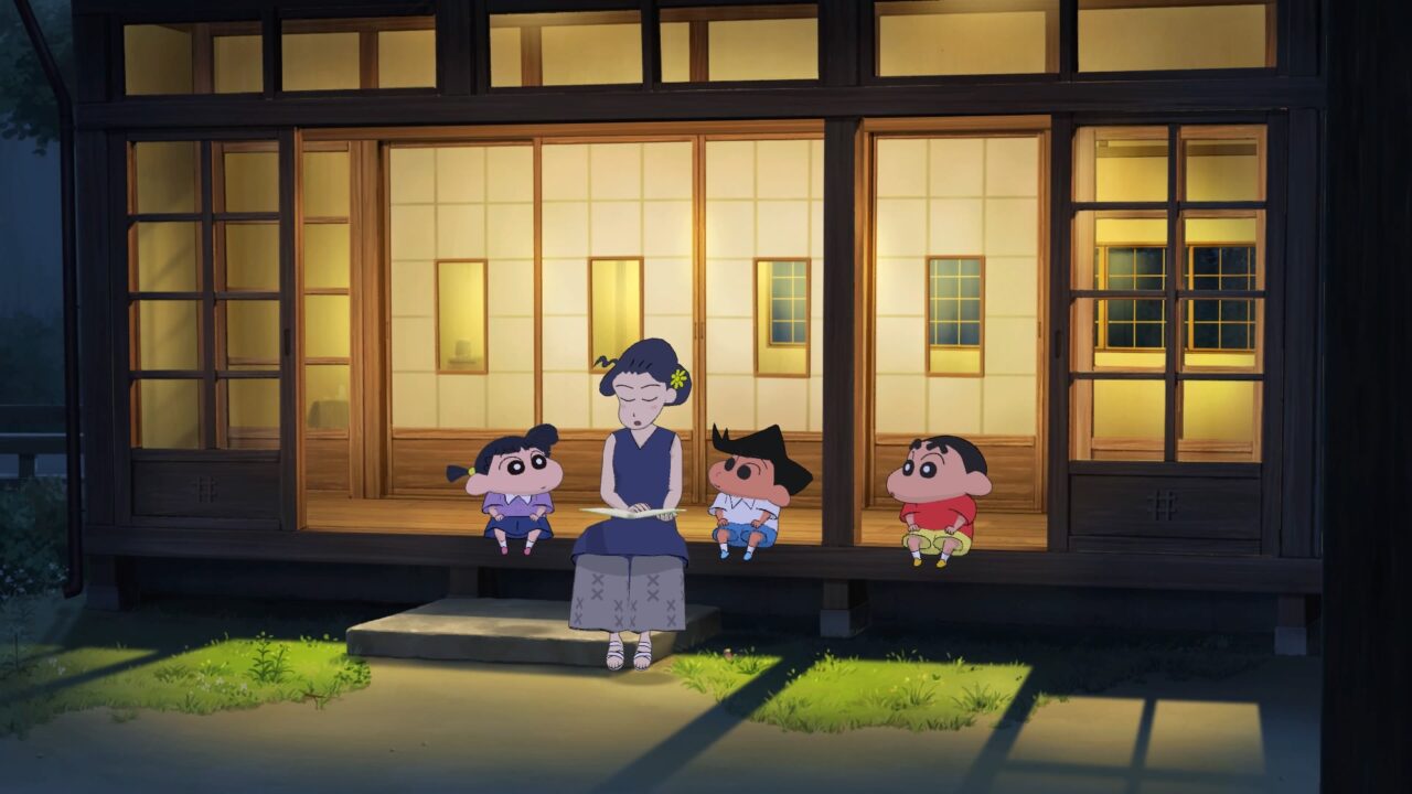 Shin Chan sitzt mit zwei anderen Kindern und einer erwachsenen Frau im Innenhof eines traditionellen japanischen Familienhauses. Die Frau liest den Kindern vor. Draußen ist es dunkel, und alle schauen sie gespannt an.