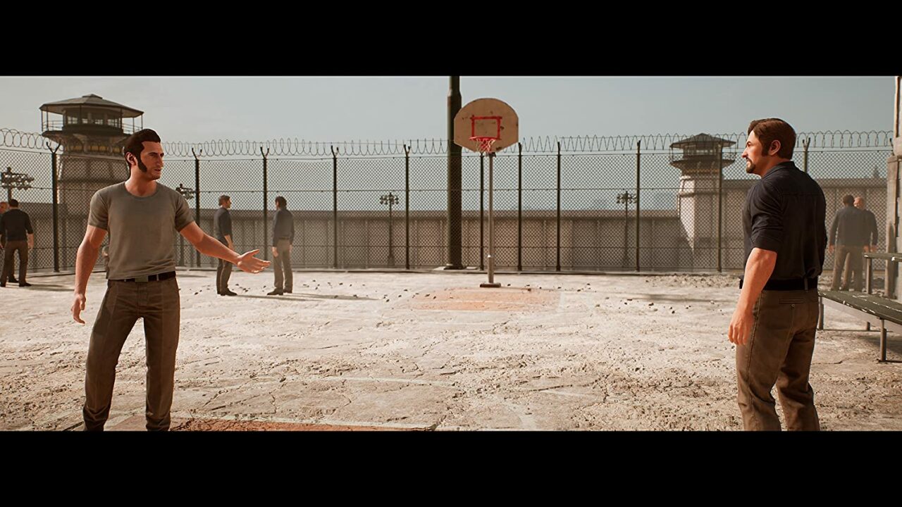 Zwei Männer befinden sich auf einem eingezäunten Sportplatz. Sie reden miteinander. Im Hintergrund stehen weitere Personen.