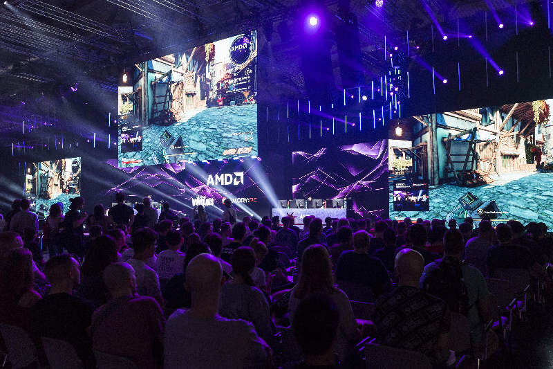 Eine Gruppe von Zuschauenden betrachtet eine Vorstellung auf der gamescom, die auf drei Leinwänden übertragen wird. Die Halle ist abgedunkelt und durch blaue, weiße und violette Lichteffekte atmosphärisch in Szene gesetzt.