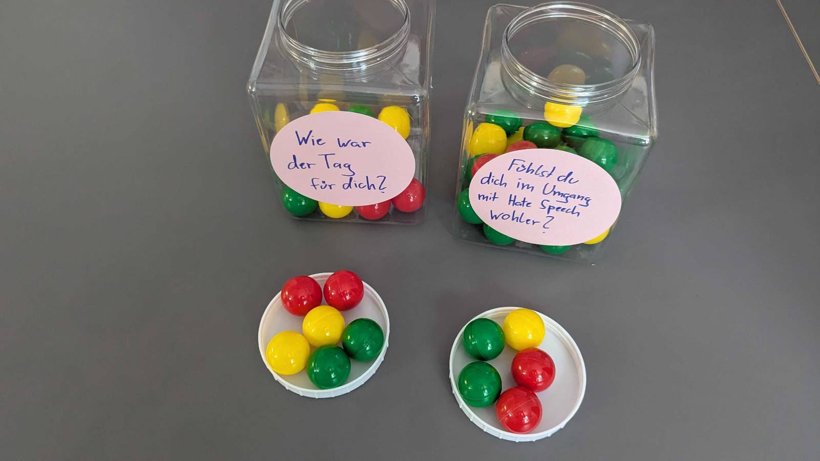 Zwei Gläser gefüllt mit roten, gelben und grünen Kugeln. Auf dem linken Glas steht: Wie war der Tag für dich? Auf dem rechten Glas steht: Fühlst du dich im Umgang mit Hate Speech wohler? Vor den Gläsern liegen weitere Kugeln.