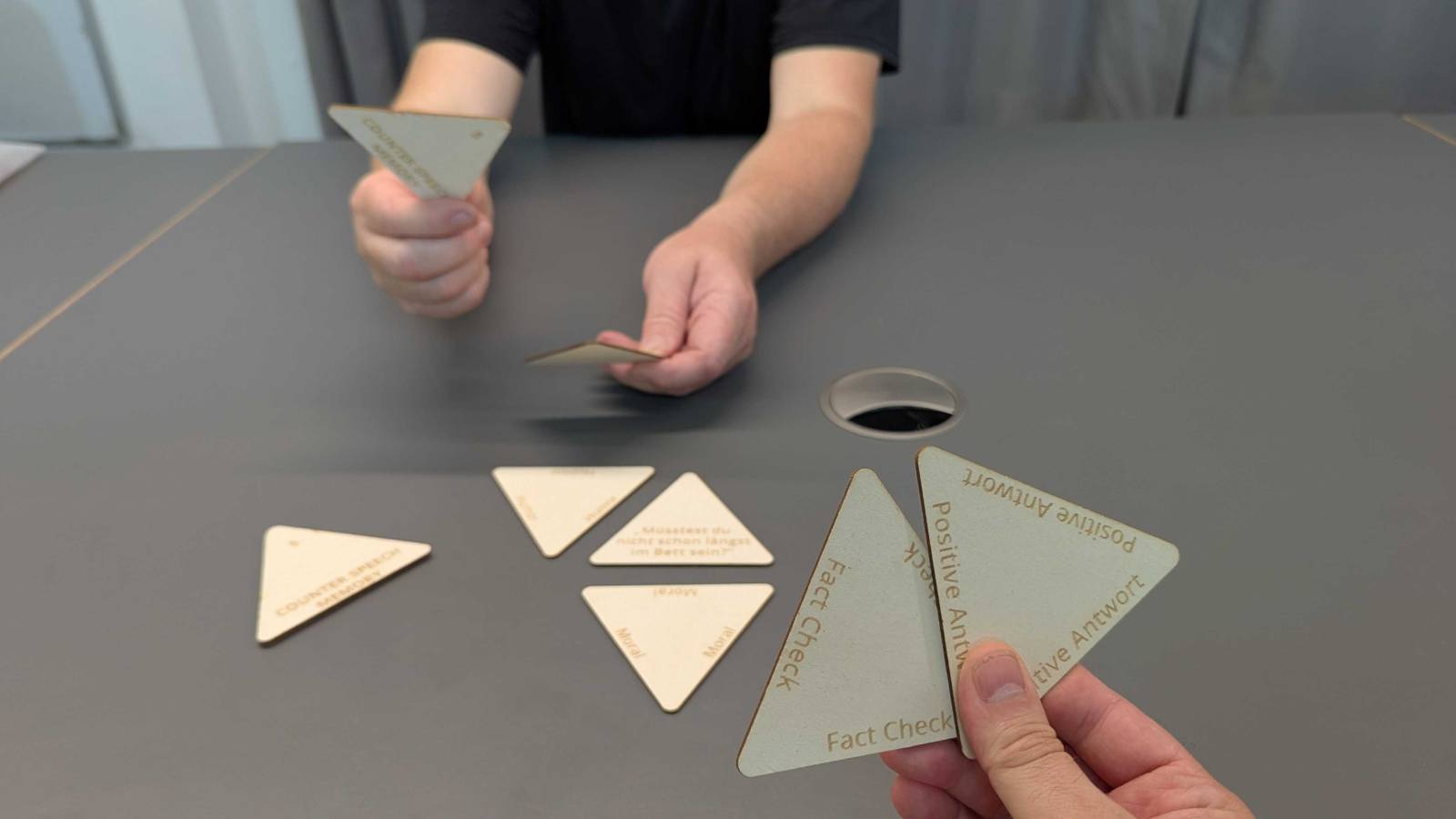 Point of View auf eine Hand mit zwei dreieckigen Spielkarten mit den Aufschriften fake Check und Positive Antwort. Gegenüber sitzt eine andere Person mit Spielkarten in der Hand. Auf dem TIsch liegen weitere Spielkarten.