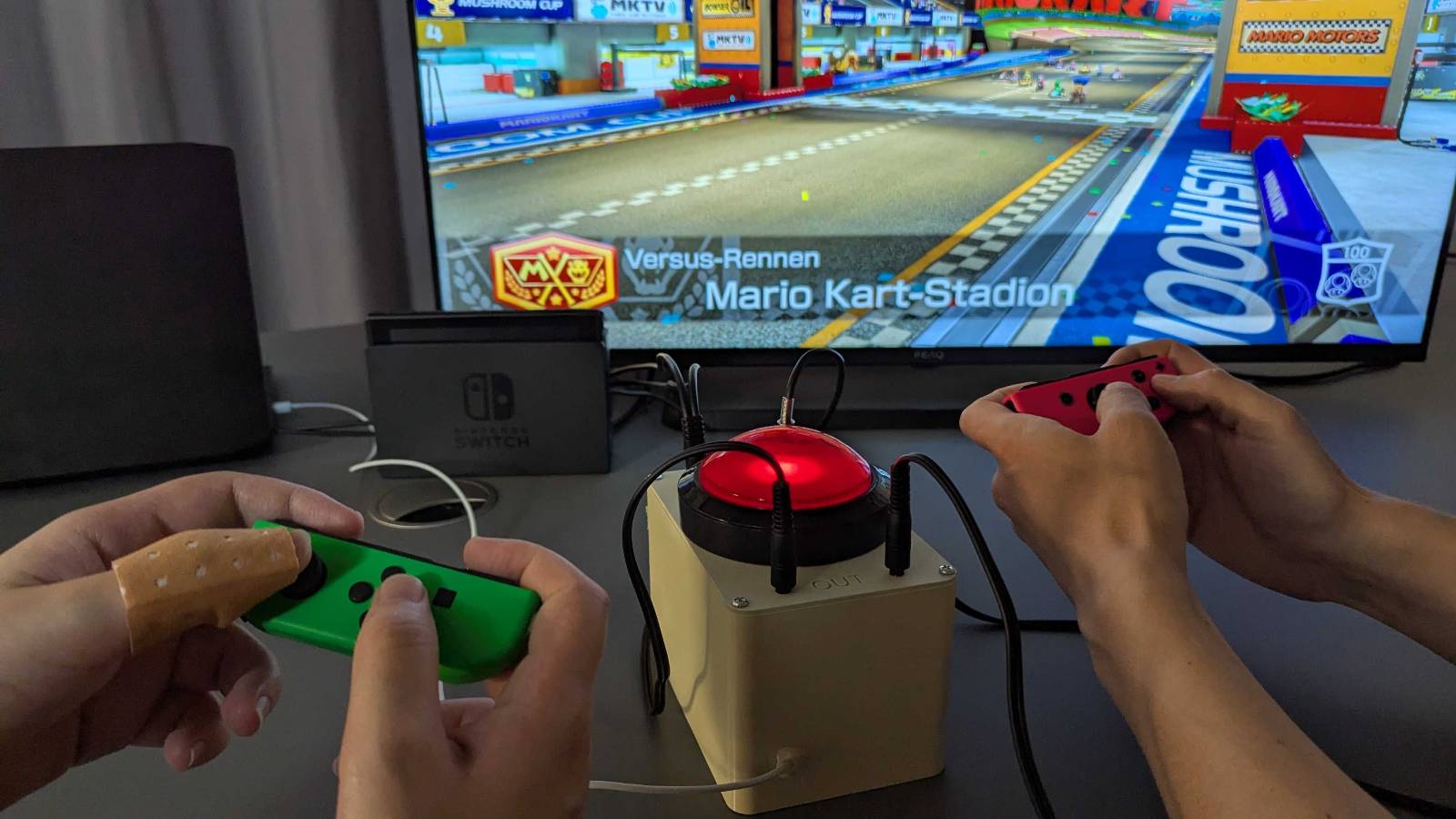 Zwei Paare Hände die jeweils einen Nintendo Switch Joycon halten. Auf einem Fernseher läuft Mario Kart 8. In der Mitte steht ein großer roter Button mit angeschlossenen Kabeln.