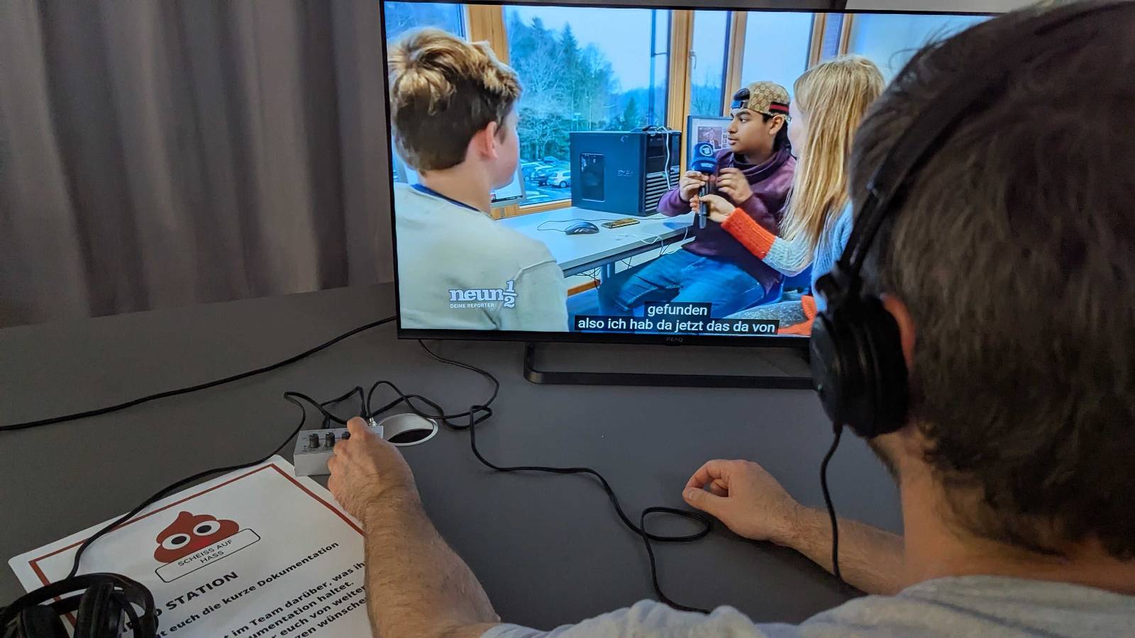 Eine Person sitzt vor einem Fernseher und hat Kopfhörer auf. Die hand der Person liegt auf einem Audiointerface mit vier Lautstärkereglern. Der Kopfhörer ist hier angeschlossen. Auf dem Bildschirm sind drei Jugendliche im Gespräch zuu sehen.