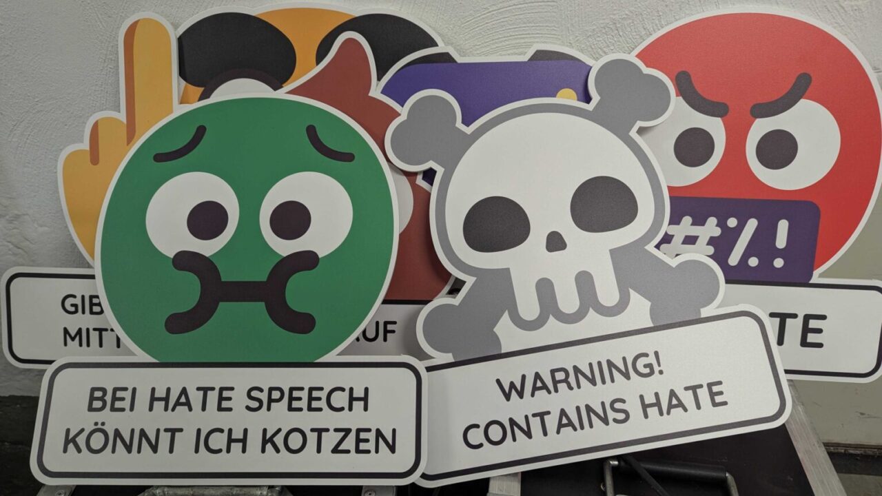 Verschiedene Emoji-Schilder übereiandergestapelt. Vorne links ein grünes Gesicht, das den Mund zukneift. Darunter: Bei Hate Speech könnt ich kotzen. Rechts vorne ein stilisierter Totenkopf. Darunter: Warning! Contains Hate.