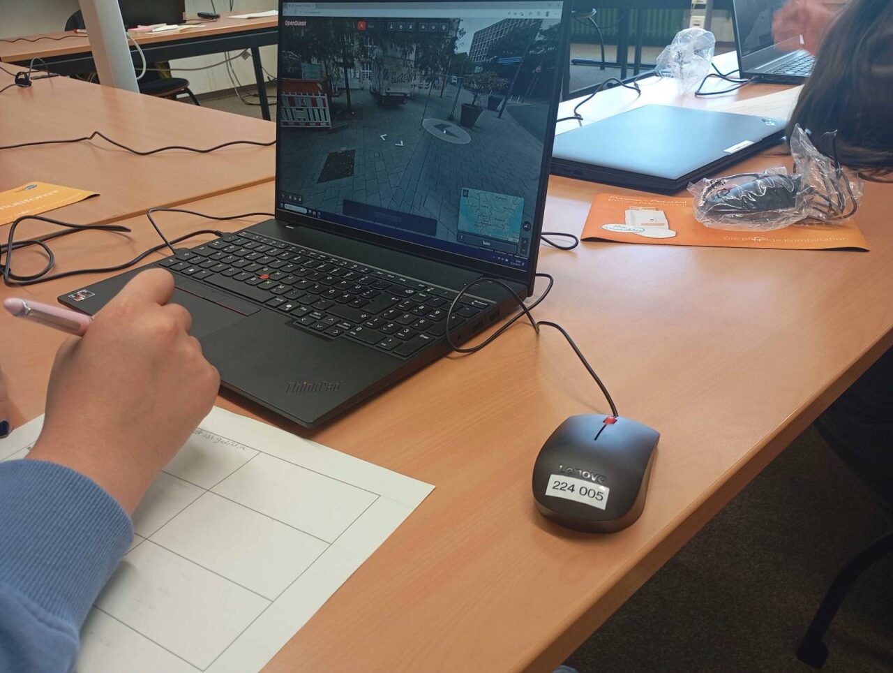 Auf einem Laptop zeigt OpenGuessr einen Ort in einer unbekannten deutschen Stadt an. Ein Jugendlicher macht sich auf einem Zettel Notizen dazu.